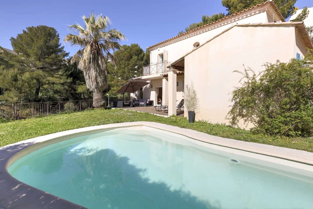 A sunny villa with a refreshing swimming pool in the foreground, surrounded by a green lawn, a large palm tree, and an outdoor patio.