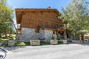 A traditional alpine chalet with a stone ground floor and wooden upper levels, featuring many shuttered windows, set among trees by a paved area.