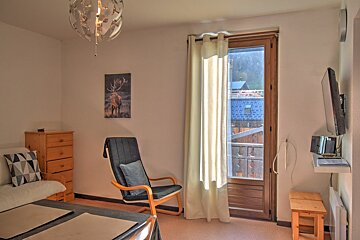 A cozy, bright room featuring a dresser, lounge chair, wall-mounted TV, and a balcony door. Decor includes deer art and a modern ceiling light.