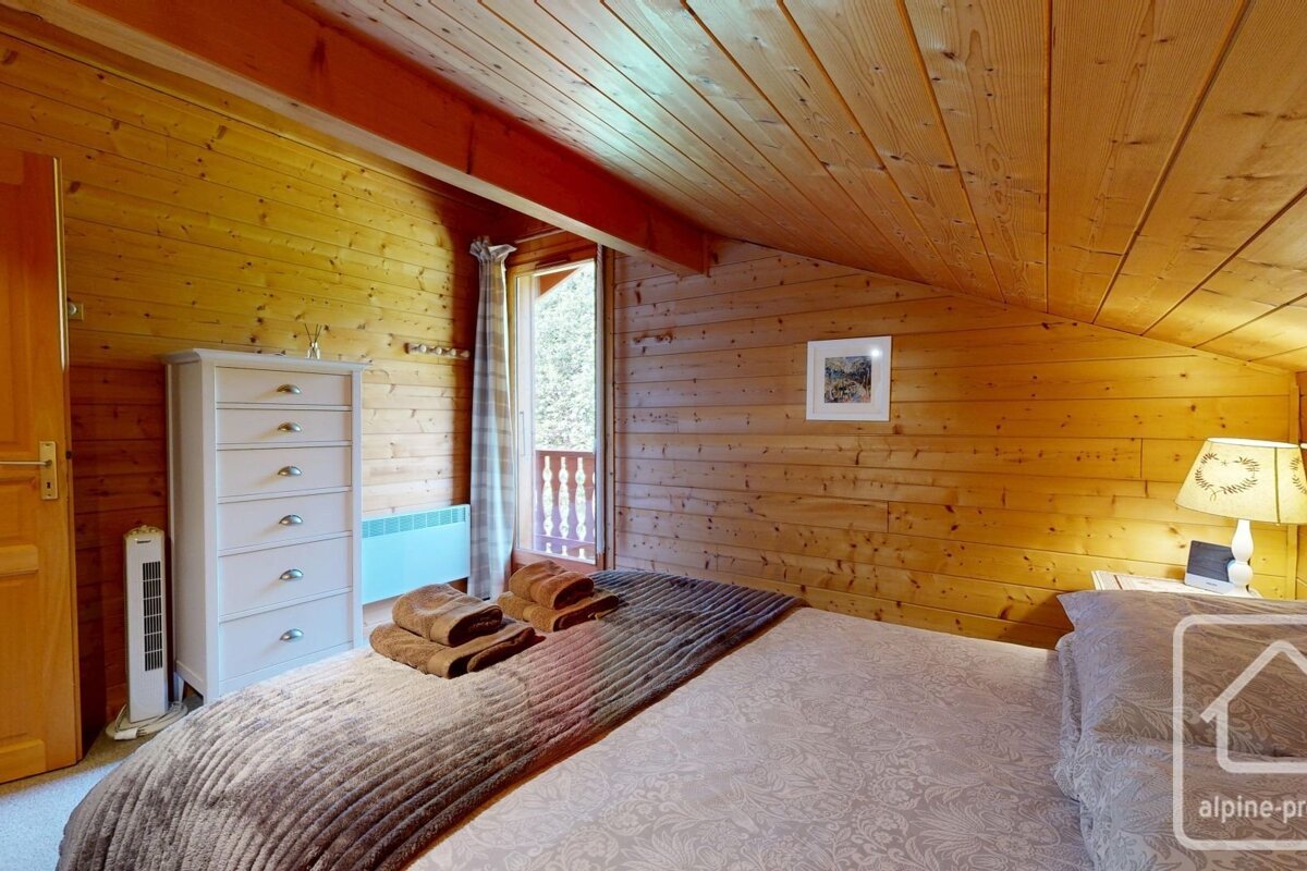 A cozy, rustic wooden bedroom features a bed, white dresser, and balcony door. A sloped wooden ceiling enhances the cabin-like ambiance.