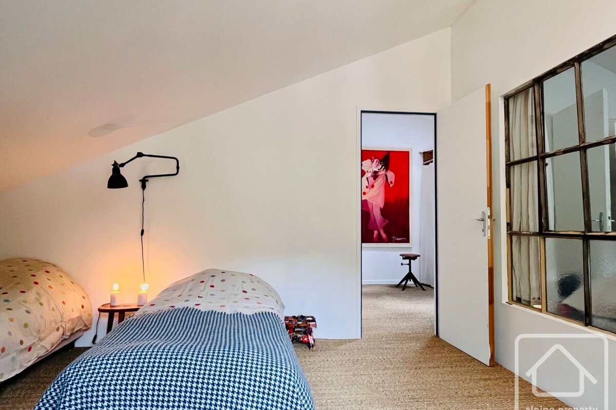 A bright, angled bedroom with two beds, warm lighting, and a doorway revealing a vibrant red painting and a stool in the adjacent room.