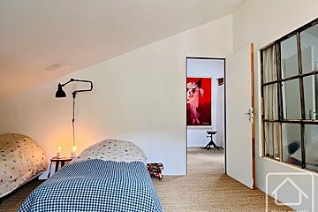 A bright, angled bedroom with two beds, warm lighting, and a doorway revealing a vibrant red painting and a stool in the adjacent room.