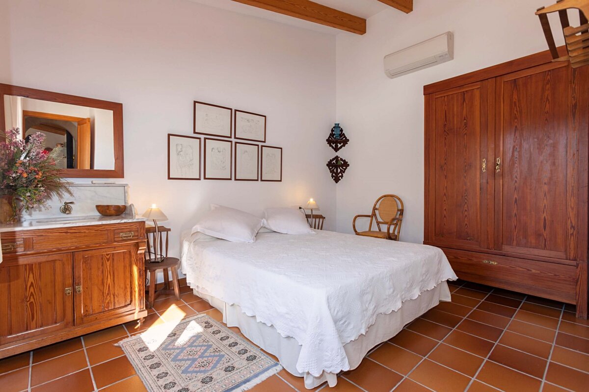 A rustic bedroom with white walls, terracotta floors, and wooden furniture including a bed with white linen, a large wardrobe, and a vanity.
