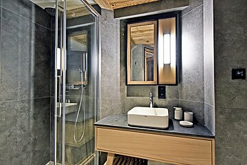 Modern bathroom with gray tiled walls, a glass shower, and a wooden vanity with a white vessel sink and a lighted mirror.