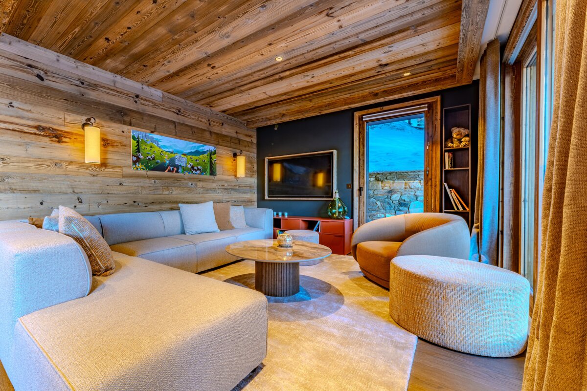 Cozy chalet living room with wood-paneled walls and ceiling, a large light-colored sectional sofa, round coffee table, and warm lighting.