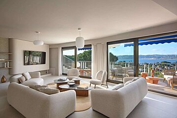 A modern, light-filled living room featuring neutral-toned furniture and large glass doors opening to a balcony with a panoramic ocean view.