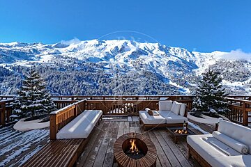 Cozy wooden deck with white outdoor seating and a fire pit, overlooking majestic snow-covered mountains under a bright blue sky.