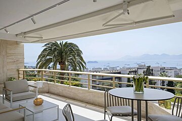 A modern balcony with stylish outdoor furniture offers a panoramic sea view, distant boats, city buildings, and a prominent palm tree under a bright sky.