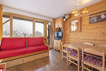 Cozy chalet living room with wood paneling, a red sofa, dining area, and large windows revealing snowy mountain village views.