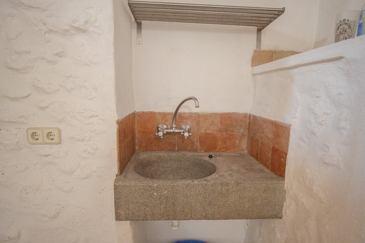 A rustic stone utility sink with a wall-mounted faucet, terracotta tile backsplash, white textured walls, power outlets, and a metal shelf.