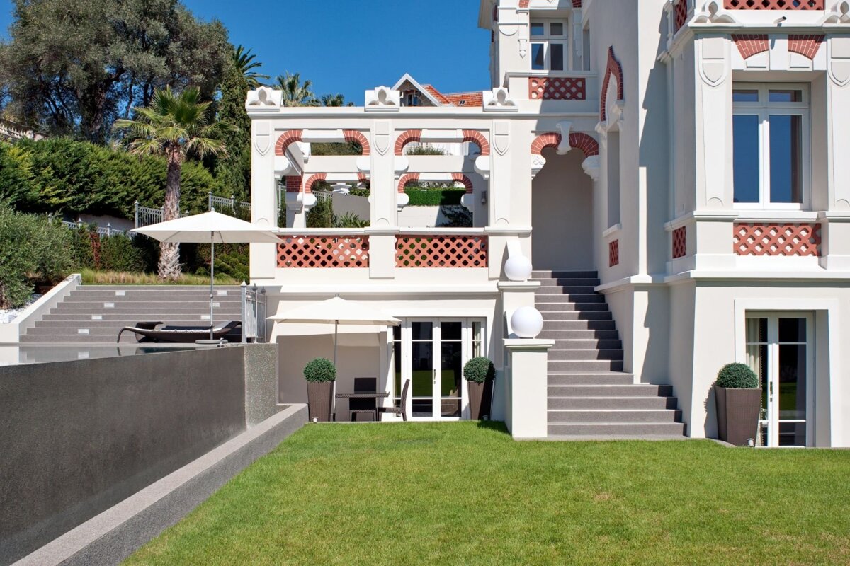 A large, elegant white villa with brick accents, featuring a green lawn, patios, steps, and outdoor seating on a sunny day.
