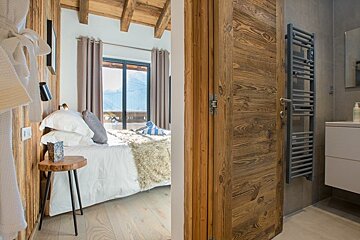 A view looking into a cozy, rustic cabin bedroom next to a modern bathroom with a wooden door.