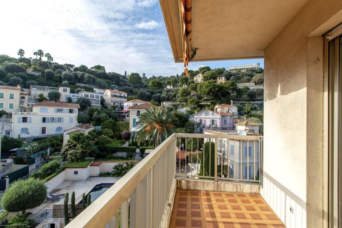 View from an apartment balcony overlooking a lush green hillside dotted with Mediterranean villas under a bright sky.