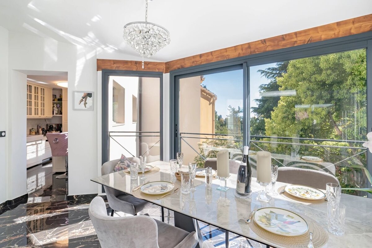 Elegant dining room featuring a set table, plush gray chairs, large windows with garden views, and a crystal chandelier.
