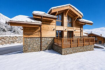 A wooden mountain chalet with stone foundations and a snow-covered roof, set in a winter landscape under a clear blue sky.