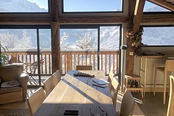 A cozy, sunlit room with wooden beams and a long dining table, offering expansive views of snow-covered mountains and trees through large windows.