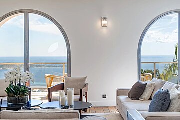 Modern living room with arched windows overlooking a stunning ocean view. Neutral decor features sofas, an armchair, and a white orchid plant.