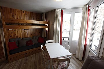 A cozy room with wood-paneled walls, featuring a bunk bed over a sofa, a white dining table, and a large window with red and white curtains overlooking a snowy scene.
