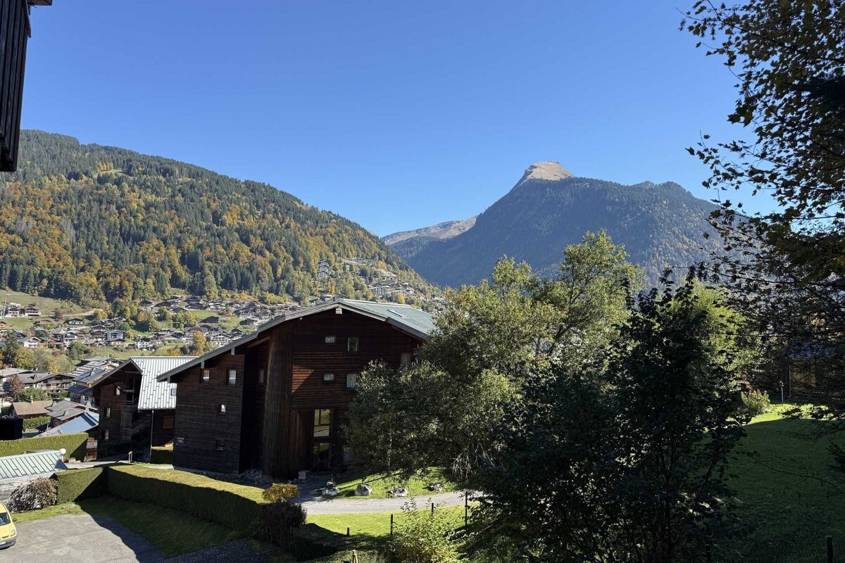 A mountain village with wooden chalets nestles in a valley, surrounded by autumn-colored forests and majestic peaks under a clear blue sky.