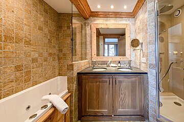 A warm, rustic bathroom featuring earth-toned tiles, wooden beams & vanity, a double sink, jacuzzi tub, and glass shower.