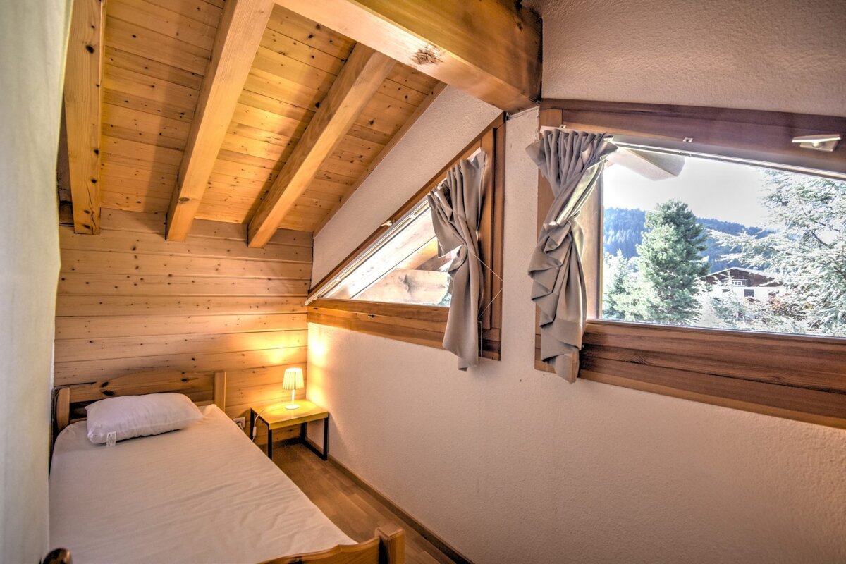 Cozy attic bedroom with a single bed, wooden walls and ceiling, and angled windows offering a scenic mountain view.