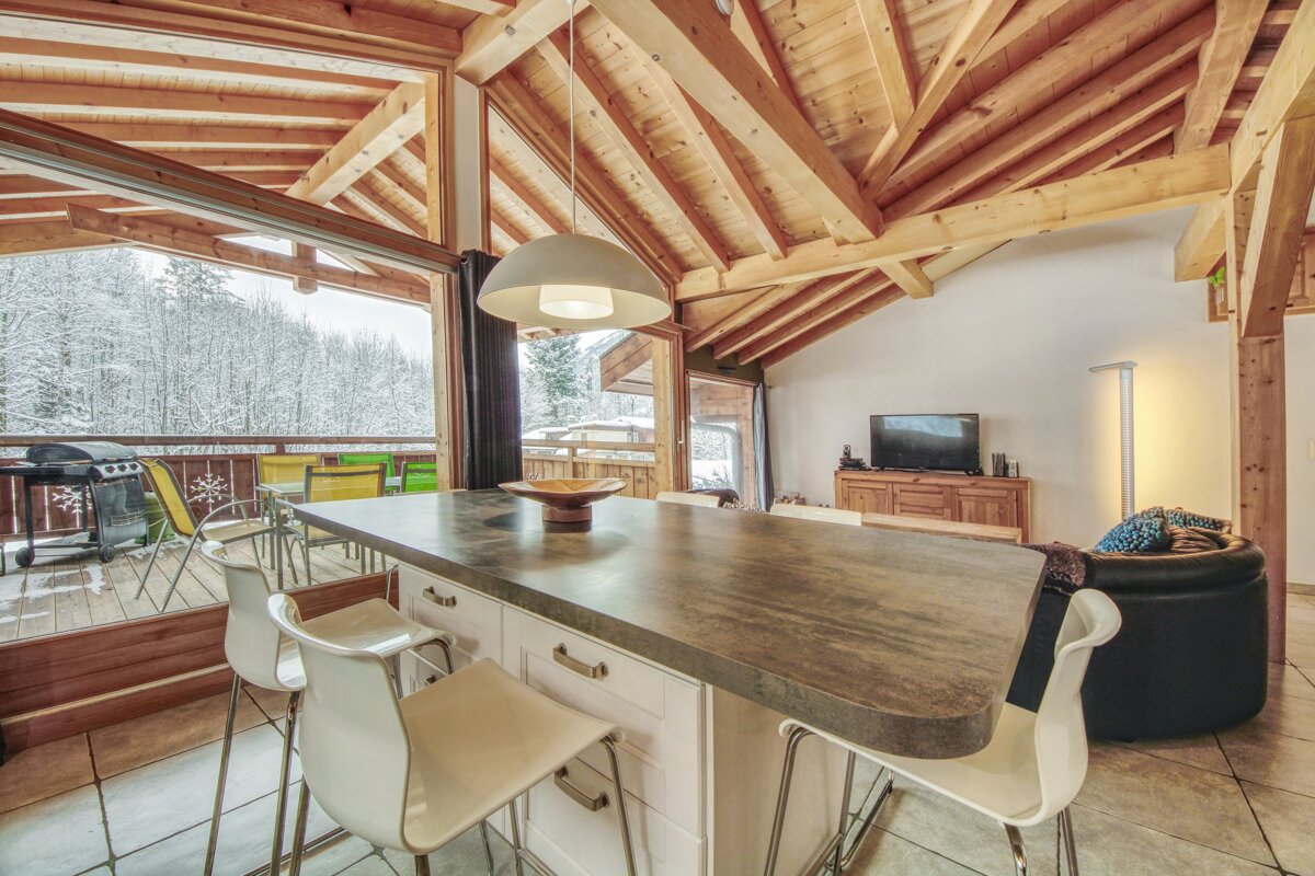 An open-plan chalet with a wooden beamed ceiling, a large kitchen island with bar stools, and panoramic views of a snowy forest from expansive windows.