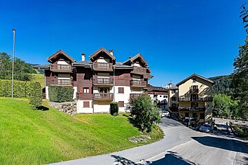 Alpine chalets with wooden balconies on a green slope next to a winding road under a bright blue sky.