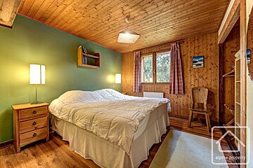 Cozy bedroom with a large bed, green and wood-paneled walls, a wooden ceiling, and rustic furniture. Plaid curtains adorn the window.
