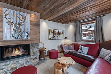 Cozy alpine living room with wood ceiling, stone fireplace, red leather sofa, wood stump tables, and deer artwork.