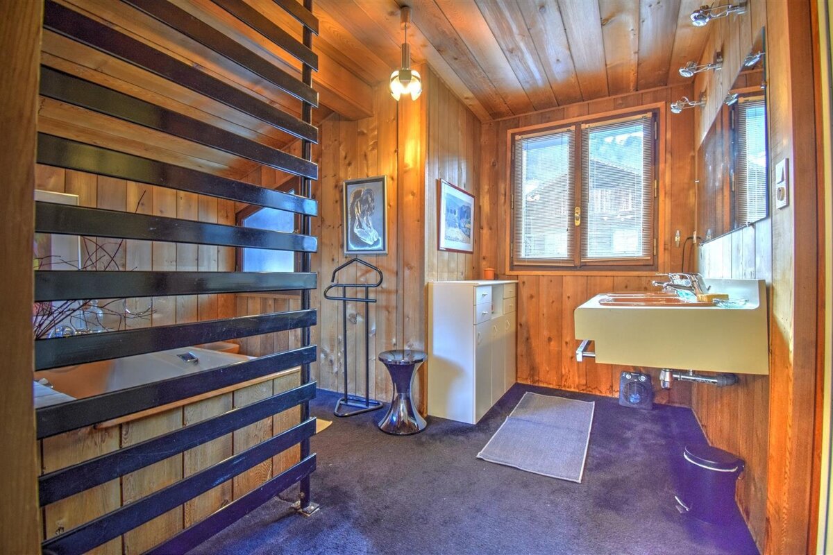 Rustic wood-paneled bathroom with a slatted partition, large sink, window, and dark carpet, featuring a vintage chalet style.