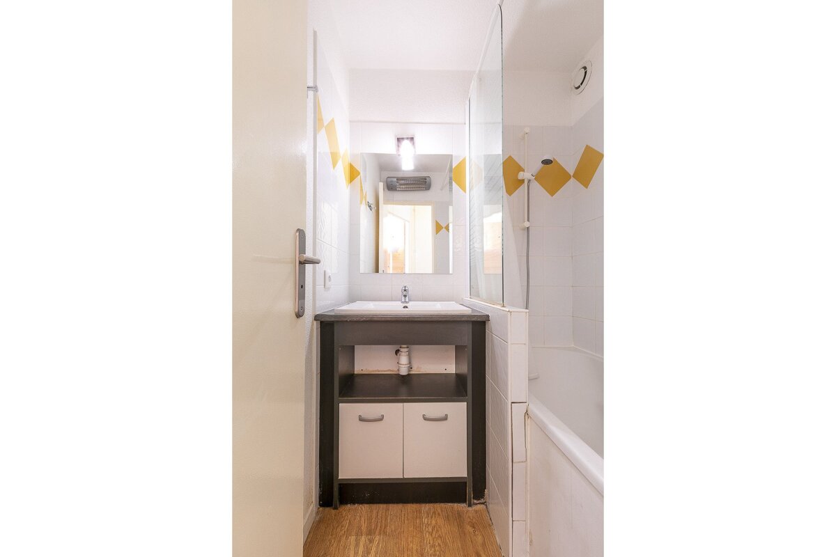 A compact bathroom features a sink vanity, mirror, and a tiled bathtub/shower combo with yellow diamond accents. A wooden floor completes the look.