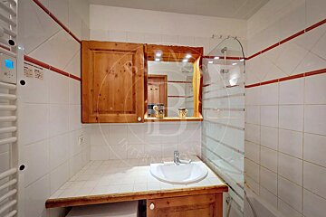 A white tiled bathroom with red border accents, featuring a wooden vanity, sink, mirror cabinet, and a shower with a glass screen.