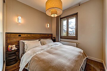 Cozy modern-rustic bedroom with a large bed, wooden headboard, neutral tones, and a window framing a scenic mountain view.