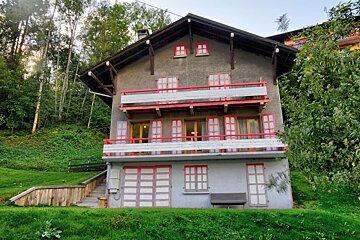 A gray multi-story house with red-trimmed balconies and shutters, nestled on a lush green hillside surrounded by trees under a bright sky.
