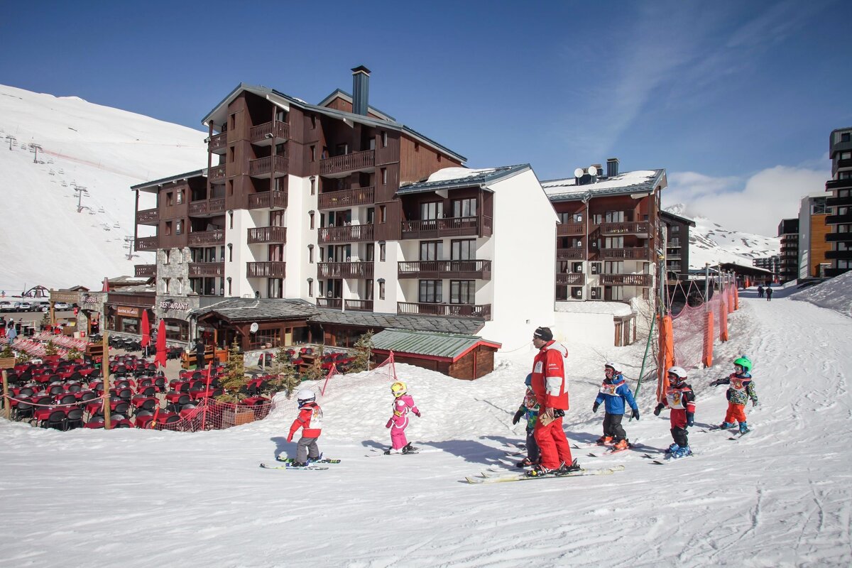 Young children learn to ski with an instructor on a sunny slope at a mountain resort, featuring large chalet-style buildings and an outdoor dining area.