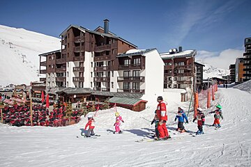 Young children learn to ski with an instructor on a sunny slope at a mountain resort, featuring large chalet-style buildings and an outdoor dining area.