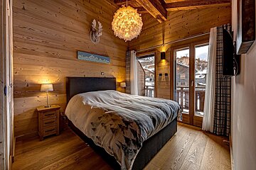 Cozy chalet bedroom with wood paneling, a large bed, fluffy chandelier, and a balcony overlooking a snowy mountain village.