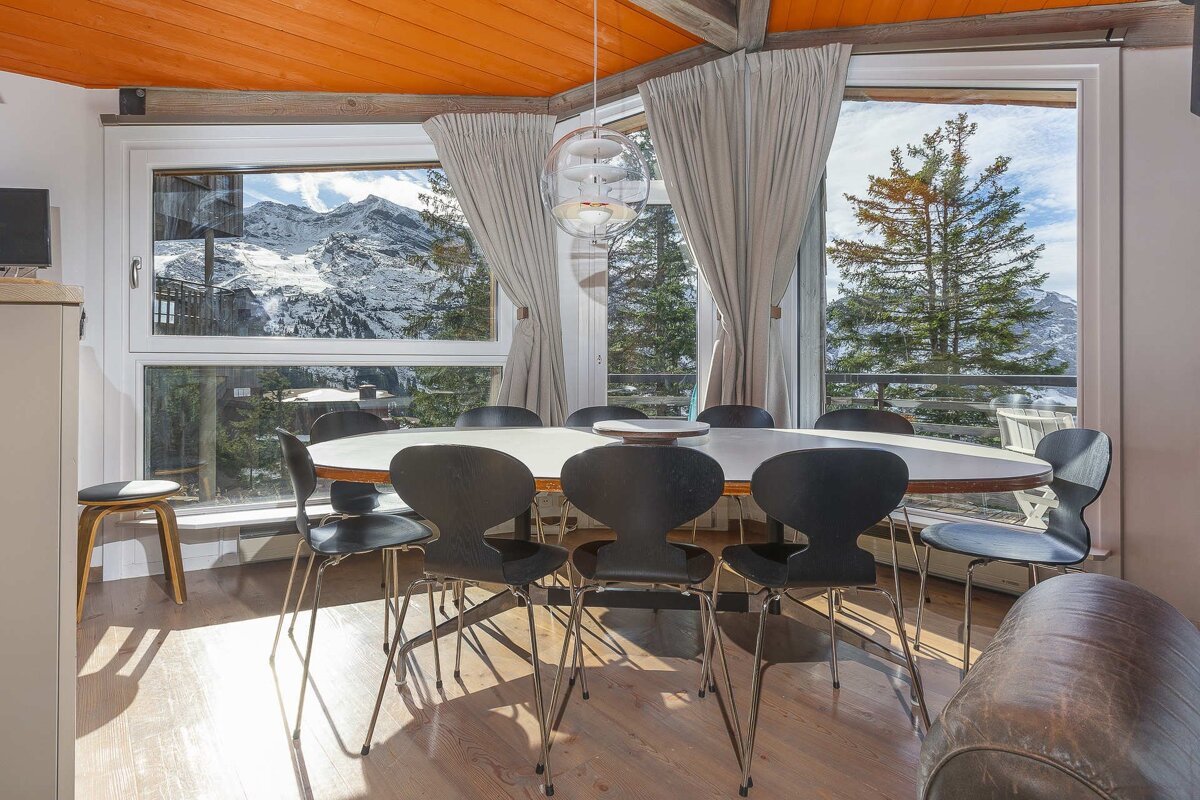 Indoor dining area with modern chairs and a white table, set against large windows revealing a breathtaking snowy mountain and pine tree landscape.