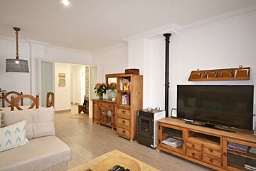 A cozy, bright living room features light wood furniture, a large TV, a pellet stove, and a dining area visible in the background.