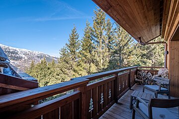 A cozy wooden chalet balcony with outdoor seating and firewood, offering a view of pine forests and snow-capped mountains under a clear blue sky.