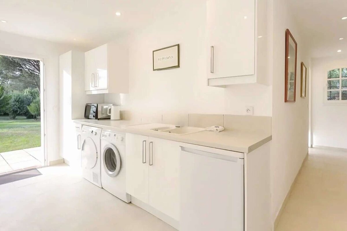 Bright, modern white utility room with a washer, dryer, sink, and cabinets. A door opens to a green garden.