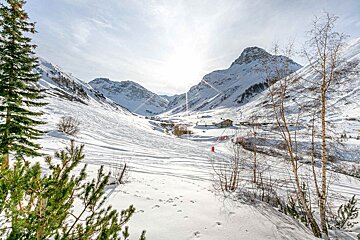 A dazzling, snow-covered mountain valley on a sunny day. Ski tracks crisscross the slopes, with scattered trees and distant buildings visible.
