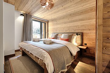 A cozy, rustic bedroom featuring extensive wooden paneling on walls, ceiling, and floor. A large bed with neutral bedding and orange accents is visible, beside a large window.