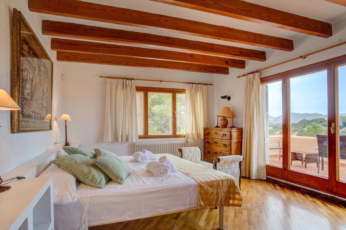A bright bedroom with exposed wooden beams, a large bed, and glass doors leading to a balcony with a scenic mountain view.