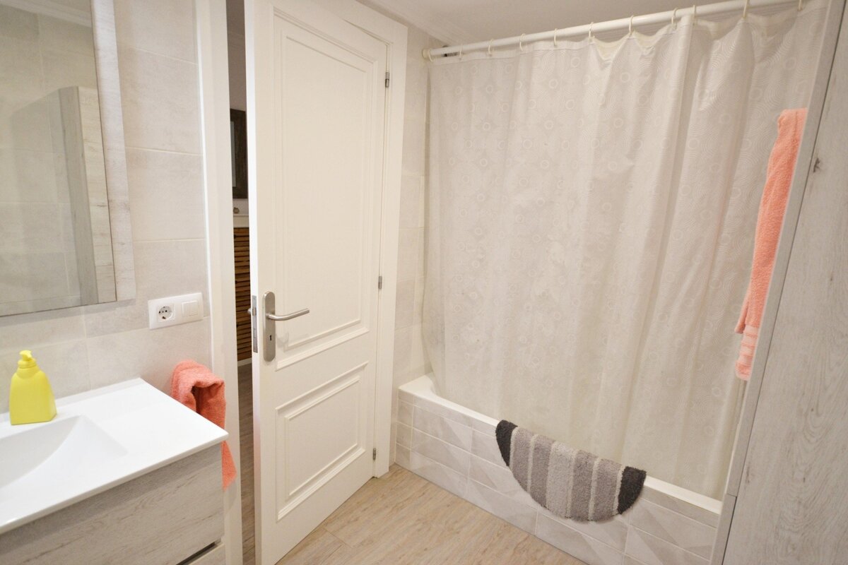 A clean bathroom with a vanity, mirror, partially open door, and a bathtub with a white shower curtain. An orange towel and striped bath mat add color.