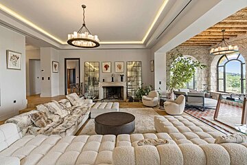 A spacious, luxurious living room with a large cream sectional sofa, fireplace, modern chandelier, and stone walls with arched windows revealing a scenic view.
