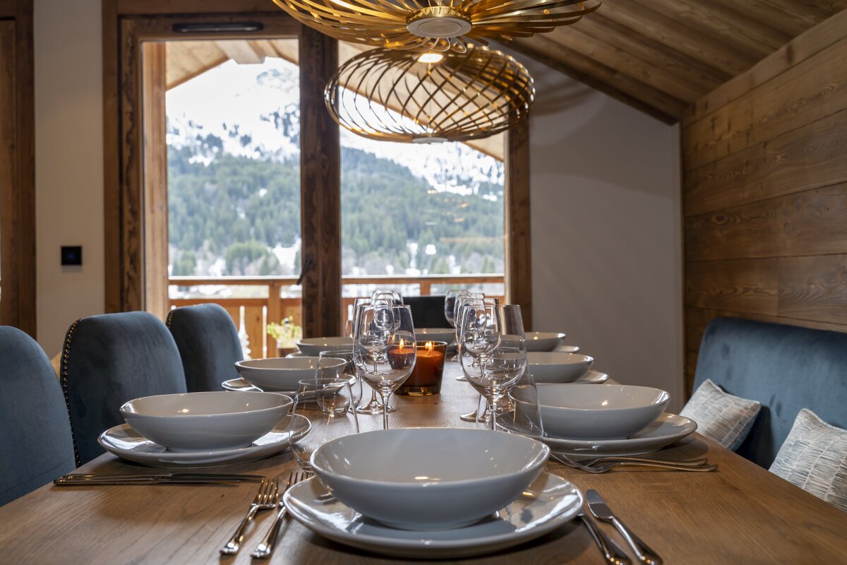 An elegantly set dining table inside a cozy wooden chalet with scenic views of snowy mountains in the background.