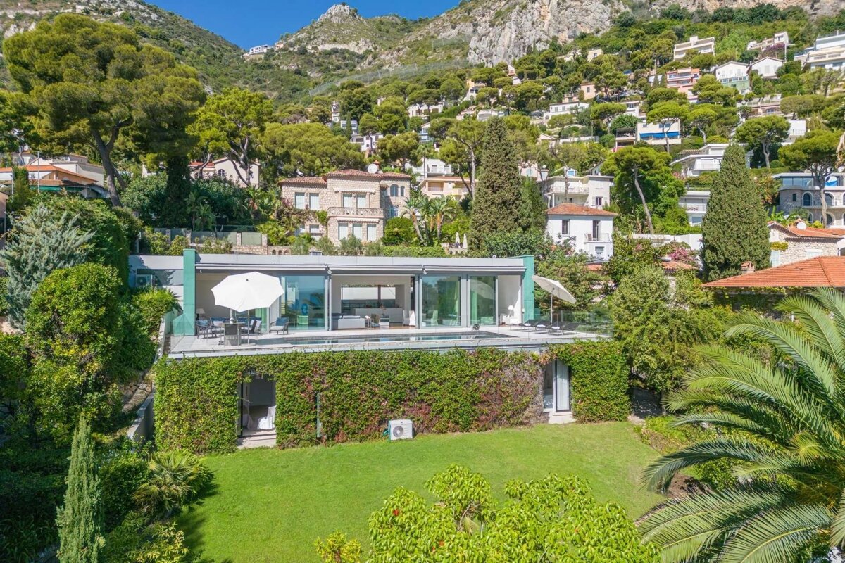 A sleek modern glass-fronted house with a terrace and pool, nestled in a green landscape with a vine-covered lower level, backed by a hillside of homes.