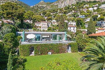 A sleek modern glass-fronted house with a terrace and pool, nestled in a green landscape with a vine-covered lower level, backed by a hillside of homes.