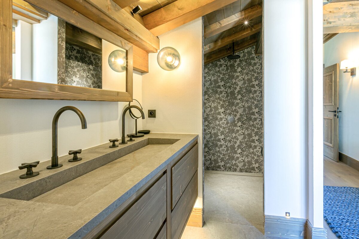 A stylish bathroom with a concrete double vanity, wooden beams, and a framed mirror, featuring a shower with floral wallpaper and warm lighting.